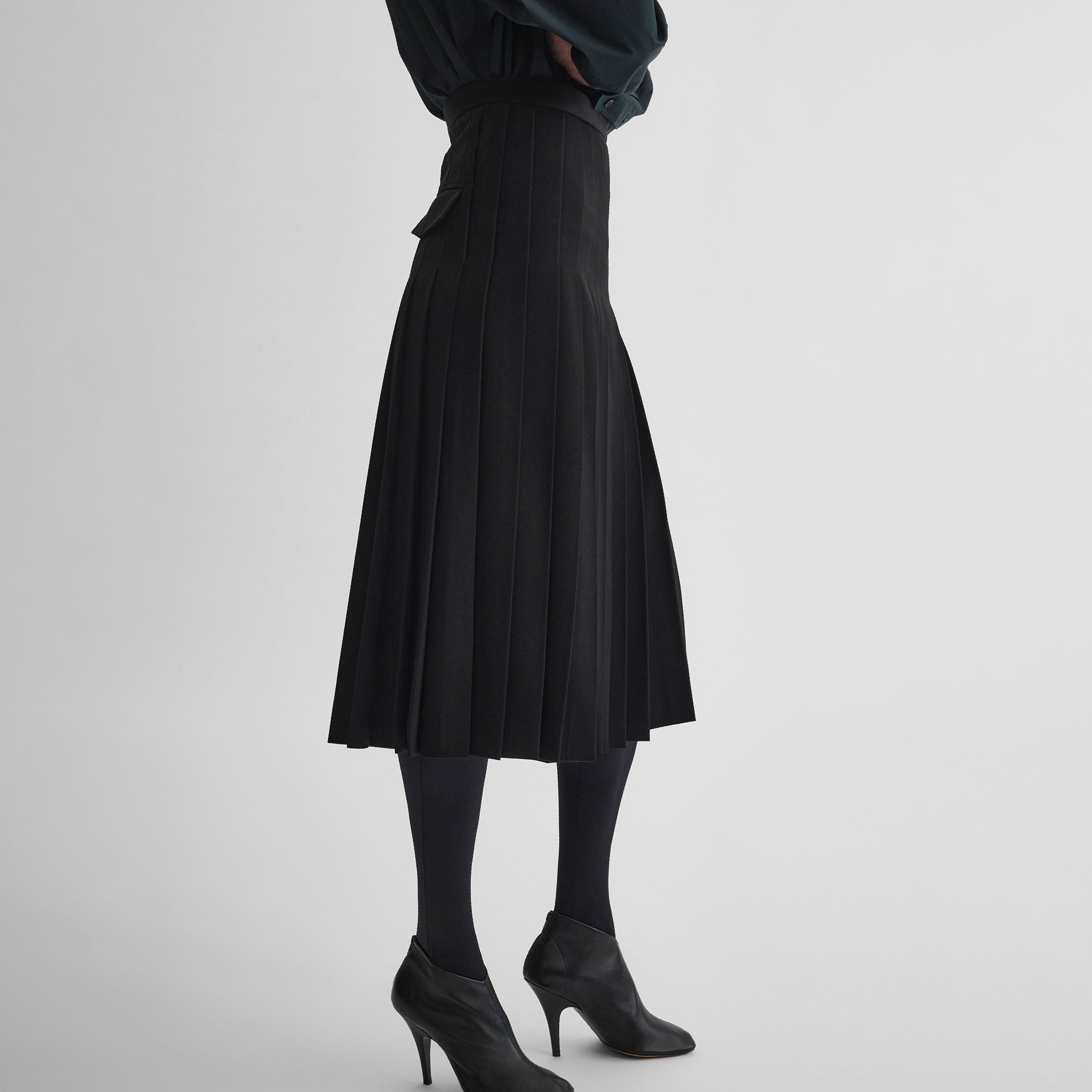 Side view of model wearing UNIFORM PLEATED SKIRT in black wool blend